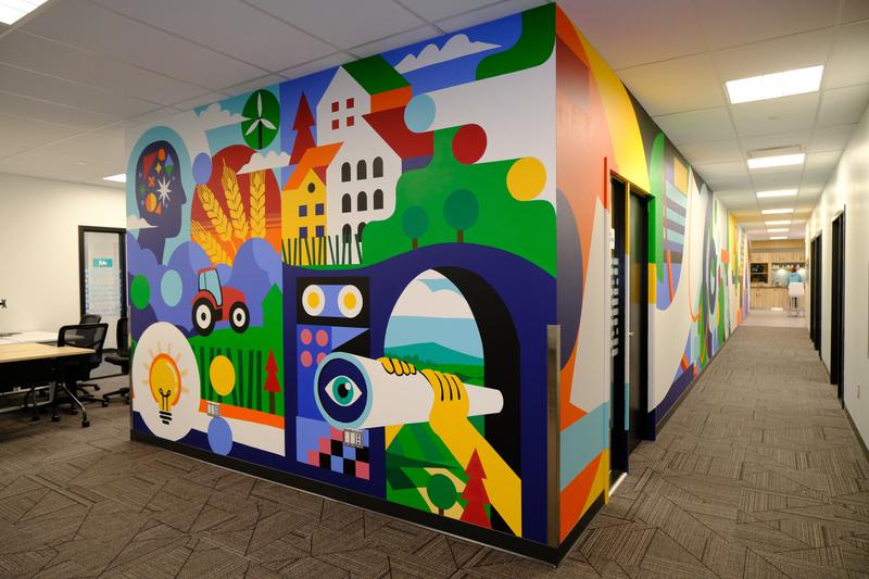 Interior office mural at Co.Labs in Saskatoon, showing a prairie scene with wheat, a tractor and a lightbulb wrapping a corner wall, with the corridor mural beyond.